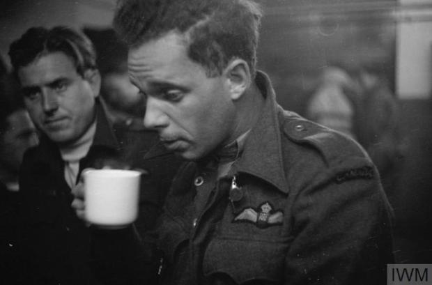 Awaiting his debrief after returning from an operation over Germany, a tired and drawn Canadian airman from No. 405 (Vancouver) Squadron Royal Canadian Air Force raises a mug of tea or coffee to his lips. His eyes appear not to be looking at anything and have a far-away look. Behind him is another colleague who looks tense. The image caption states that it was made during July or August 1942 by Pilot Officer Forward, RAF.
